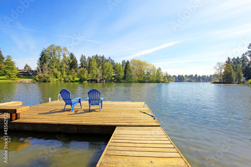 Lake waterfront with pier and two blue chairs.