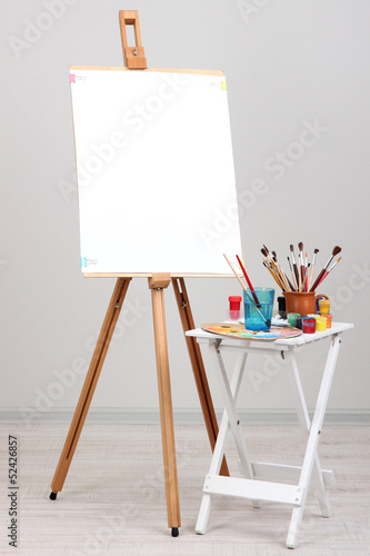 Wooden easel with clean paper and art supplies in room