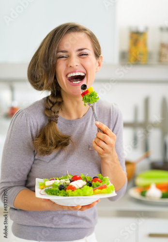 Happy young woman eating greek salad in kitchen