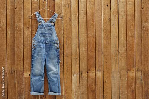 Pair of denim dungarees hanging against wooden wall.