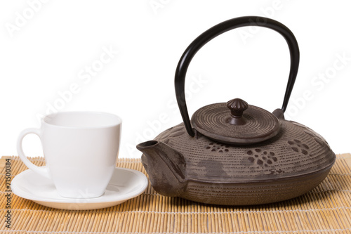 White cup of tea on saucer and iron teapot on wooden mat.