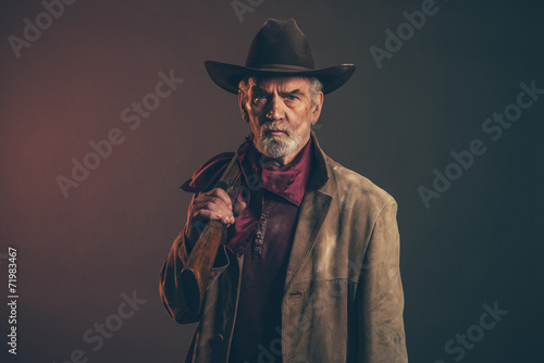 Old rough western cowboy with gray beard and brown hat holding r