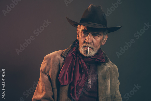 Old rough western cowboy with gray beard and brown hat smoking a