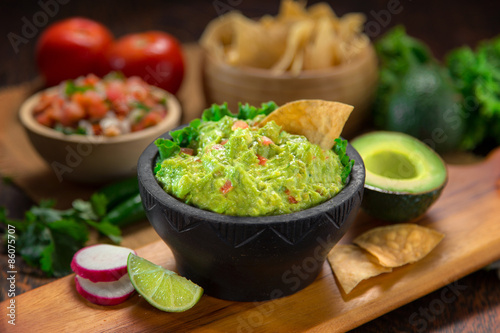 A delicious Bowl of Guacamole next to fresh ingredients on a table with tortilla chips and salsa