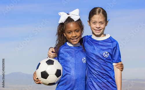 Cute, young african american and hispanic female soccer players holding a ball with a simple blue sky background