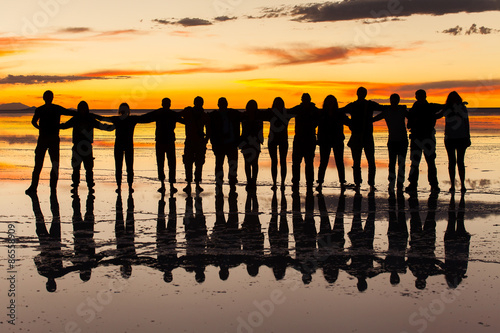 Group of friends embraced in the sunset light and wet salt surface in salares de uyuni, Bolivia. Orange sky with clouds background