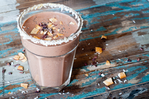 Chocolate coconut smoothie milkshake blended with cacau, coconut milk, banana and topped with coconut flakes and chia seeds. A healthy snack or treat served in a tall glass on a rustic wooden table.