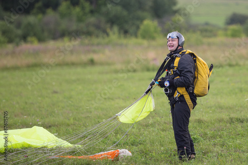Joyful man in black suit with yellow parachute standing on green field after jump