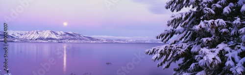 To jezioro Tahoe po śnieżnej burzy śnieżnej. Nad jeziorem unosi się księżyc w pełni, a drzewa i góry są pokryte śniegiem.