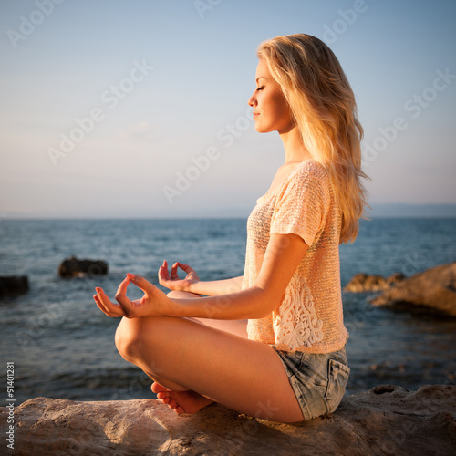 Beautiful young blond woman meditating on a beach at sunrise in