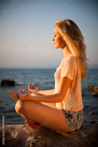 Beautiful young blond woman meditating on a beach at sunrise in