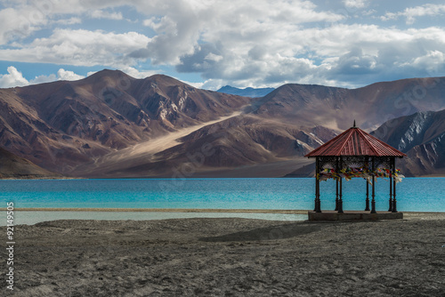  Kashmir Pavilion with pray flag stand alone at Pangong lake with beauty view surrounded by mountain range and turquoise colour lake. 