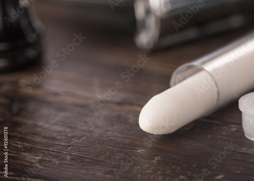 Styptic pencil in clear tube in bathroom setting on wooden counter with double edged razor and brush handle in the background