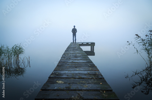 Man standing on a jetty at a lake during a foggy, gray morning.