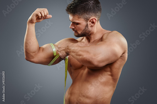Closeup handsome strong athlete measuring muscle biceps with tape measure isolated over gray background. Bodybuilder tries to measure bicep by himself.