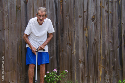 A frail one hundred year old centenarian senior citizen man with a cane standing by a wooden fence. Ample room for text in negative space over the fence.