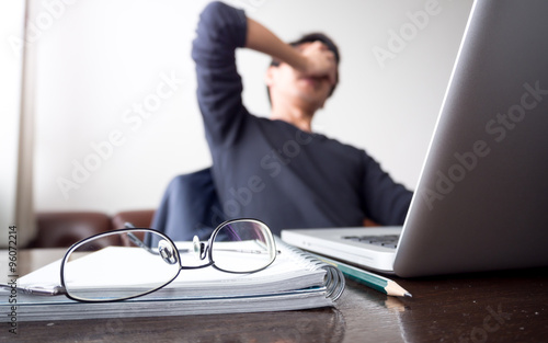 man with his hand holding his face taking a brake from working with laptop computer and notebook with eye glasses on wooden desk. concept of stress/rest/tension/failed/discourage