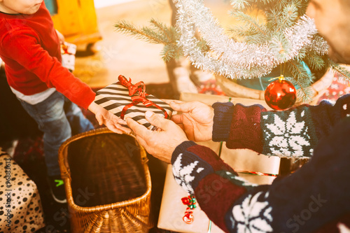 A blond haired toddler and her family exchanging Christmas gifts near the Christmas tree on Christmas Day to celebrate the event. The child takes with the hand the gift his father gives him 