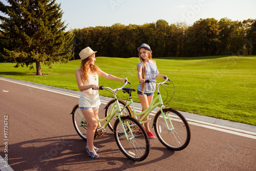 two pretty girls with a hats walking with a bicycle on park