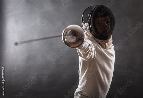 Boy wearing white fencing costume and black fencing mask standing with the sword practicing in fencing. 