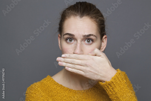 negative feelings concept - portrait of surprised beautiful 20s girl covering her mouth for bad breath or taboo,studio shot on gray background