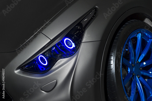 Predatory car headlight with blue lights and hood of powerful sports car with matte grey paint and wheel with blue disc 