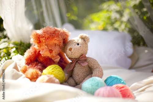 Два плюшевых медведя сидят на кровати рядом с клубками разноцветной пряжи