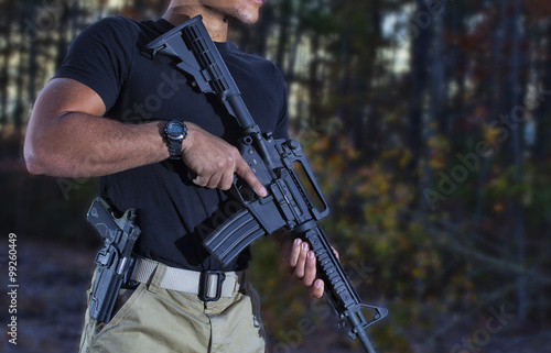 Man in a forest holding an assault rifle with handgun on his side ready to shoot