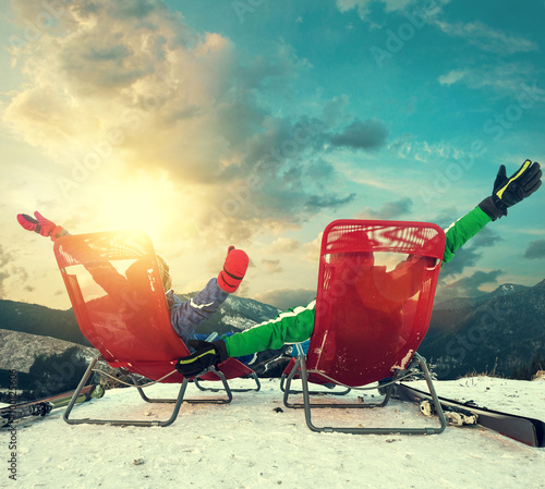 Two happy skiers sitting in chaise-longues on the top of snow mo