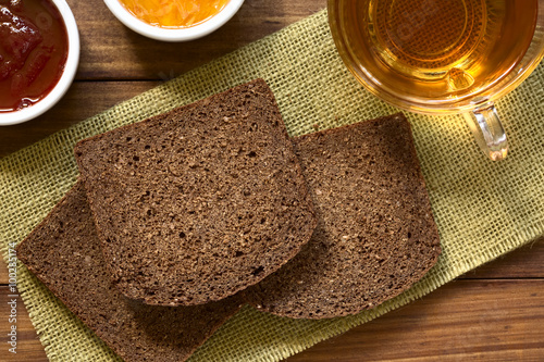 Slices of pumpernickel dark rye bread with strawberry and orange jam, cup of tea on the side, photographed overhead with natural light (Selective Focus, Focus on the upper slice)