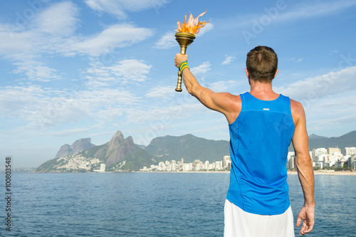 Athlete in athletic uniform standing with sport torch in front of Rio de Janeiro Brazil skyline at Ipanema Beach