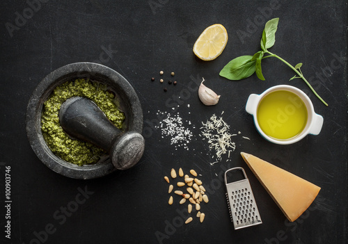 Green basil pesto - italian recipe ingredients on black chalkboard background from above. Parmesan cheese, basil leaves, pine nuts, lemon, olive oil, garlic, salt and pepper.