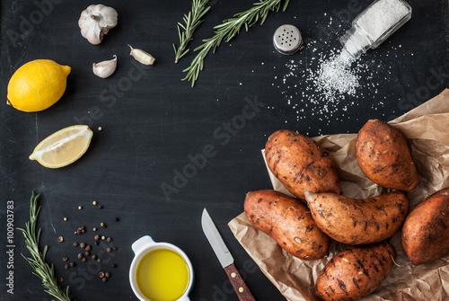 Preparing rosemary roasted sweet potatoes with olive oil, lemon, salt, pepper and garlic - kitchen scenery from above. Black chalkboard as background. Layout with free text space.