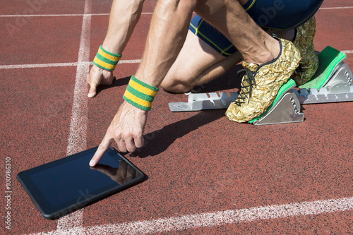 Athlete in gold running shoes crouching at the starting line of a running track wearing Brazil colors wristbands using his tablet