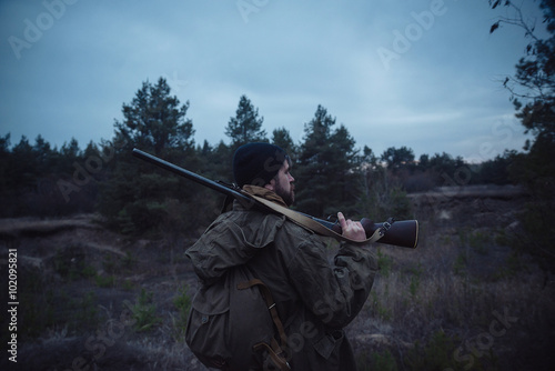 brutal hunter, bearded man in warm hat with a gun in his hand, a knife a backpack and smoking pipe in the dark wild forest in the autumn