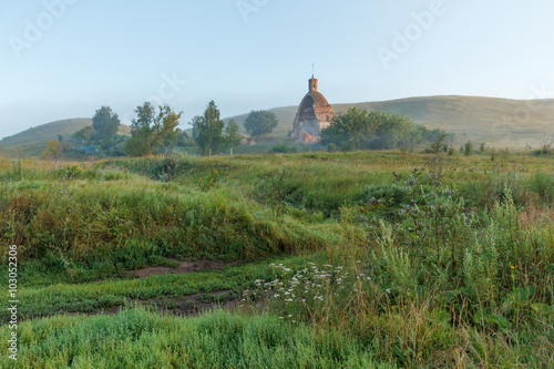 Summer landscape with dirt road and destroyed the old rural church on the hilly bank of the river a sunny misty morning