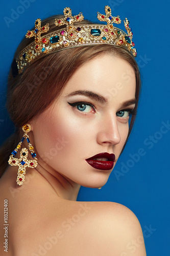 Girl with a golden crown and golden earrings. The crown with precious stones, diamond, sapphire. Earrings in the shape of a cross. Brown-haired girl. Blue background.