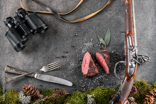 deer or venison steak with antique long gun, cutlery, binocular and ingredients like sea salt, herbs and pepper, food background for restaurant or hunting loving