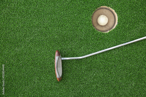 Putting golf club on green grass with golf ball in the hole - top view