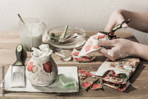 Decoupage accesories - brush, sponge, paint, tissues, pencils, scissors and papers on wooden table - hands cutting flowers in the background