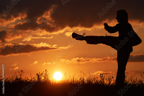 Man practicing karate on the grassy horizon at sunset. Karate kick leg. Art of self-defense. Silhouette on a background of dramatic clouds at sunset.