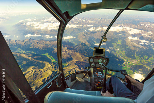 Helicopter cockpit flying on mountain landscape and cloudy sky, with pilot arm driving in cabin. Spectacular aerial view of Alps mountain chain.