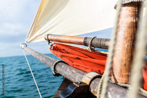 wooden bowsprit, mast, jib sail blew up by the wind and yardarm with an ocher sail and ropes at the bow of an old sailing boat with the sea at the blur background during a sunny sea trip in brittany