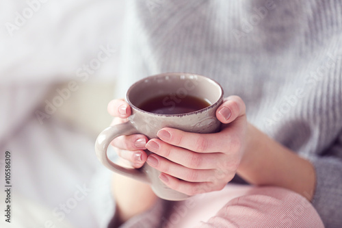 Woman in a grey sweater and warm socks holding a cup of tea while sitting on a white knitted blanket