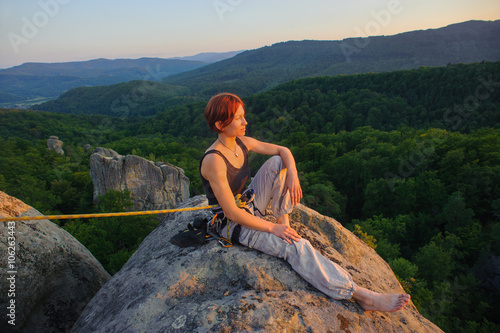 Young female rock climber sitting secured with rope on big rock at mountain peak with bare foot relaxing after climbing. Warm sunny summer evening in the mountains. Climbing equipment.