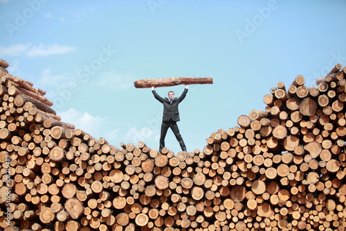 Hardworking Business Man on top of large pile of logs lifting heavy log - front view