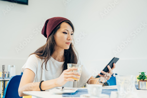 young handsome asiatic woman sitting in a bar, holding a smart phone and glass of juice, looking down and tapping the screen - technology, social network, communication concept
