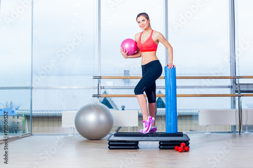 Fitness girl, wearing in sneakers, red top and black breeches, posing on step board with ball, the sport equipment background, gym