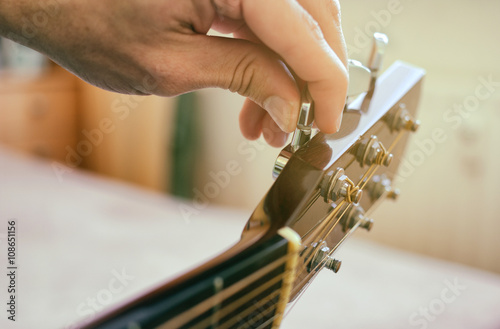 Tuning the guitar. Fingers are turning the tuning peg on the head of acoustic guitar. Authentic shot with blurred room in the background.