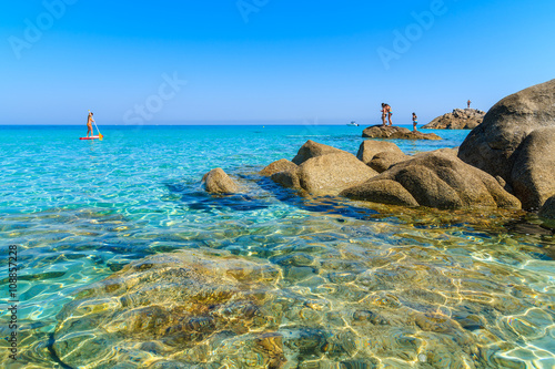 Crystal clear sea water of Bodri beach and woman swimming on surfboard in distance, Corsica island, France